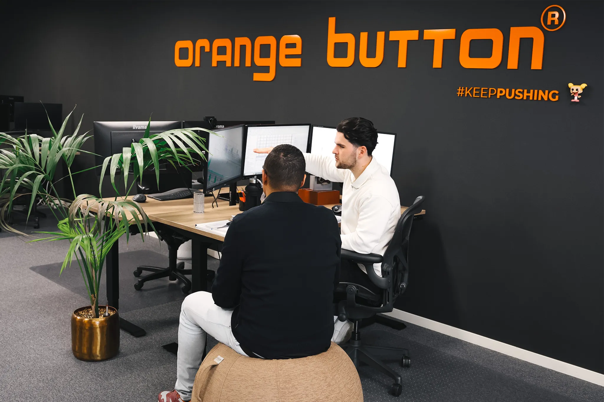 Twee collega's van Compact aan het werk achter een scherm, met het Orange Button-logo op de muur