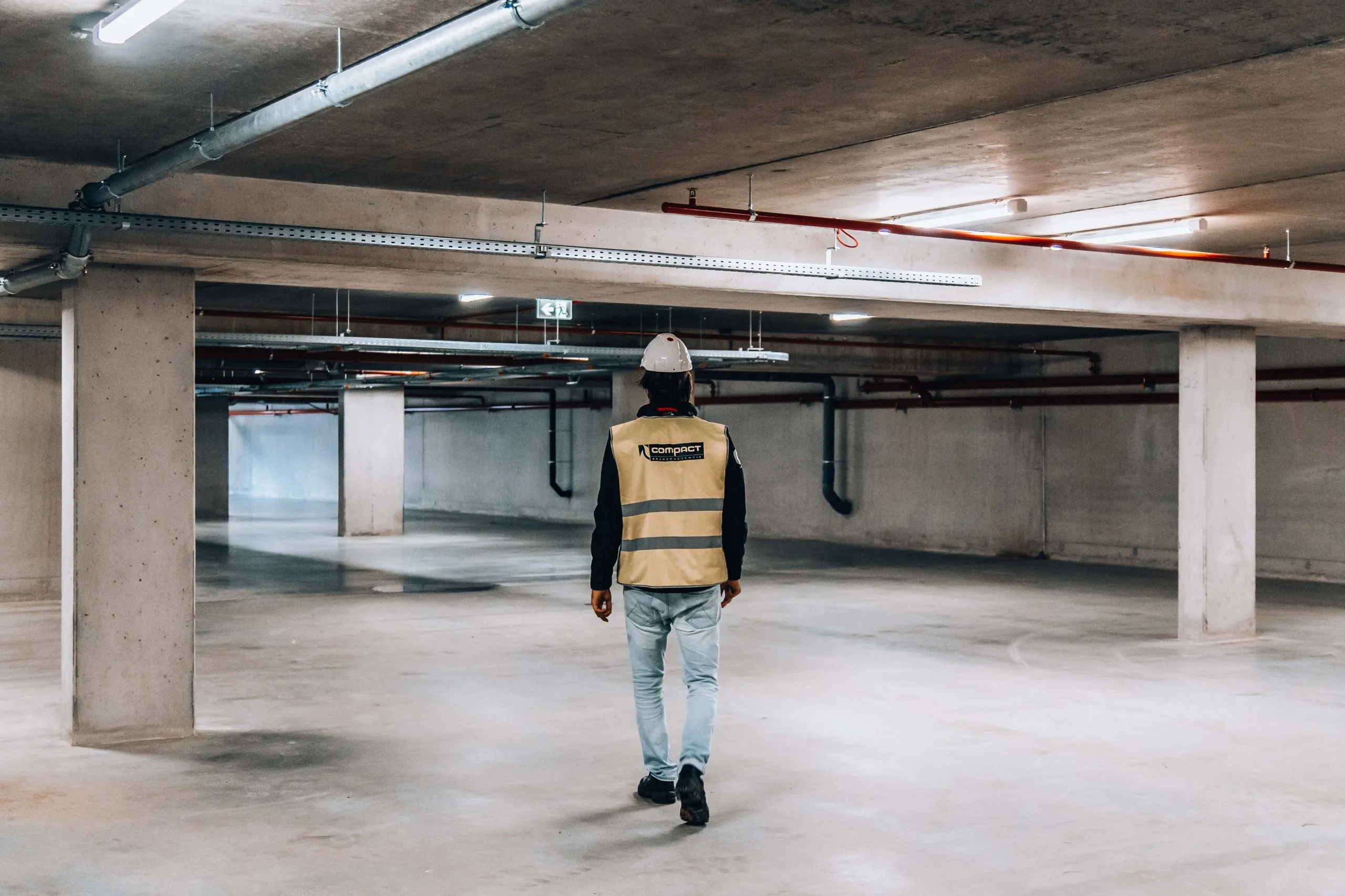 Een monteur van Compact loopt door een parkeergarage met leidingen en kabelgoten aan het plafond